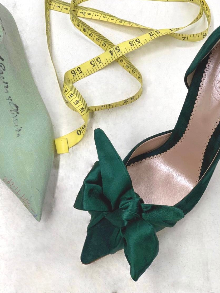 Green suede heels with a large bow detail on the front, positioned on a soft white surface. A measuring tape is placed next to the shoe, indicating a measurement context. The angle of the image showcases the shoe from above, highlighting its elegant design and the smooth interior. image