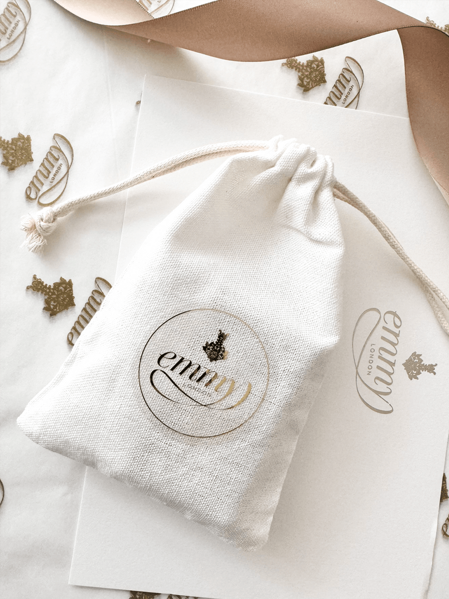 A small white drawstring bag with a gold logo is placed on a light-colored surface adorned with gold-embossed emblems. The background features soft, neutral tones, creating an elegant and minimalistic presentation. image