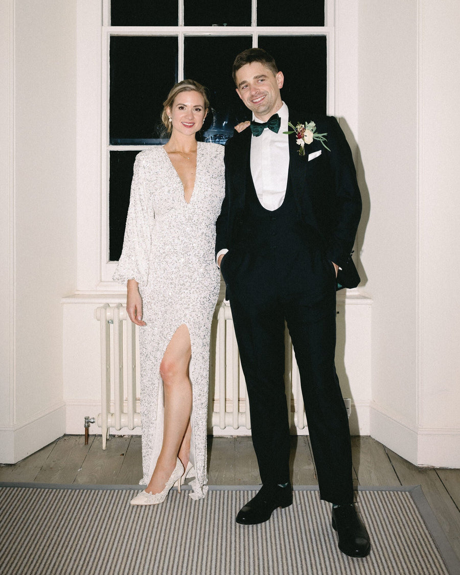 Elegant white heels made of leather, featuring a pointed toe and a modest heel height, complementing a sparkling long-sleeve gown. The model stands beside a man in a tuxedo, both posing in a well-lit room with large windows and a neutral-toned rug. image