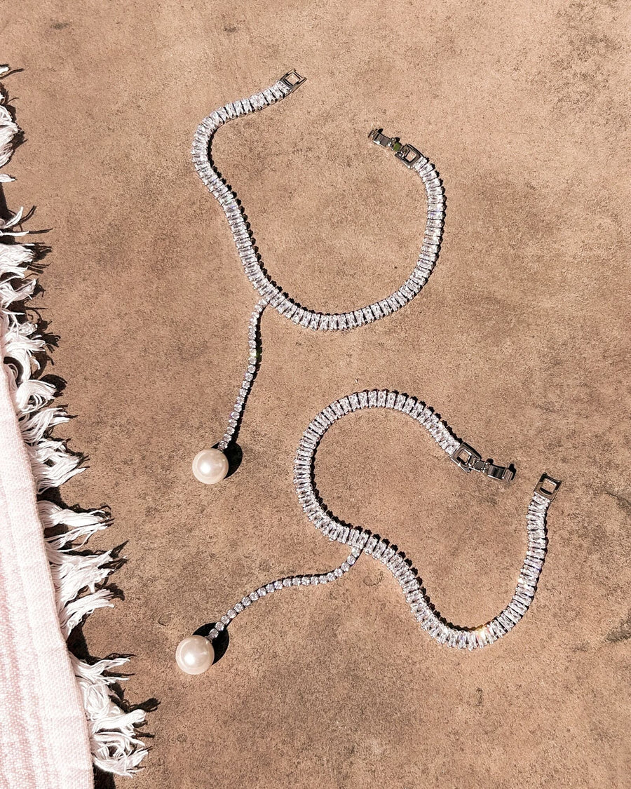 Two elegant ankle bracelets are displayed on a sandy surface. Each bracelet features a series of sparkling rhinestones, with one adorned with a single pearl drop. The background is a soft, neutral tone, enhancing the shine of the jewelry. The angle captures the bracelets from above, showcasing their intricate design and luxurious appeal. image