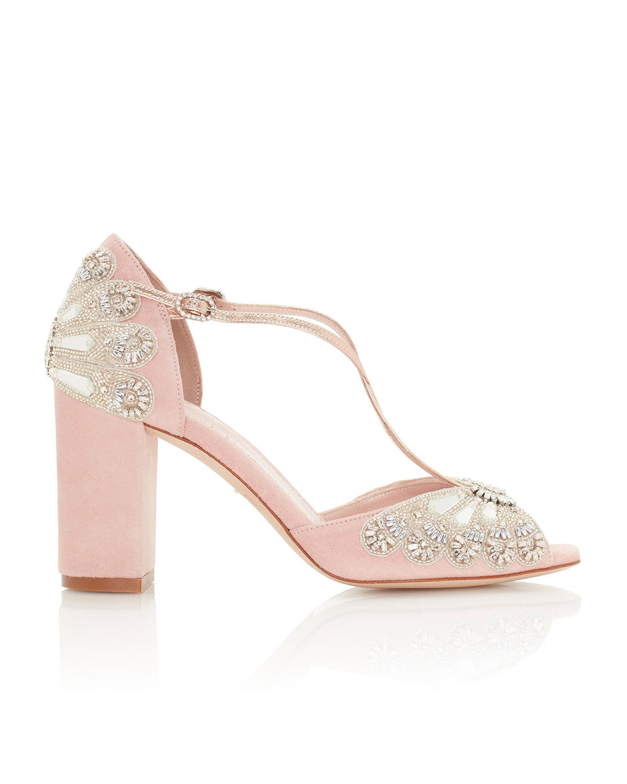 Elegant pink suede heels featuring intricate embellishments on the back and sides. The shoes have a block heel design, providing stability and comfort. The image is taken from a side angle, showcasing the detailed craftsmanship and the open-toe style. The background is a clean, white surface, emphasizing the shoes' design. image