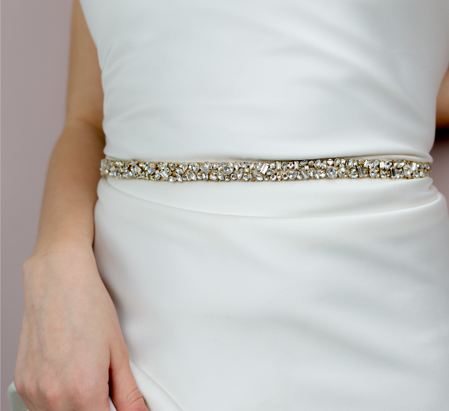 A close-up image of a woman wearing a white dress with a fitted silhouette. The dress features a sparkling gold and crystal belt that accentuates the waist. The background is a soft, muted color, enhancing the elegance of the outfit. The woman's hand is gently resting on the belt, showcasing the intricate details of the embellishments. image