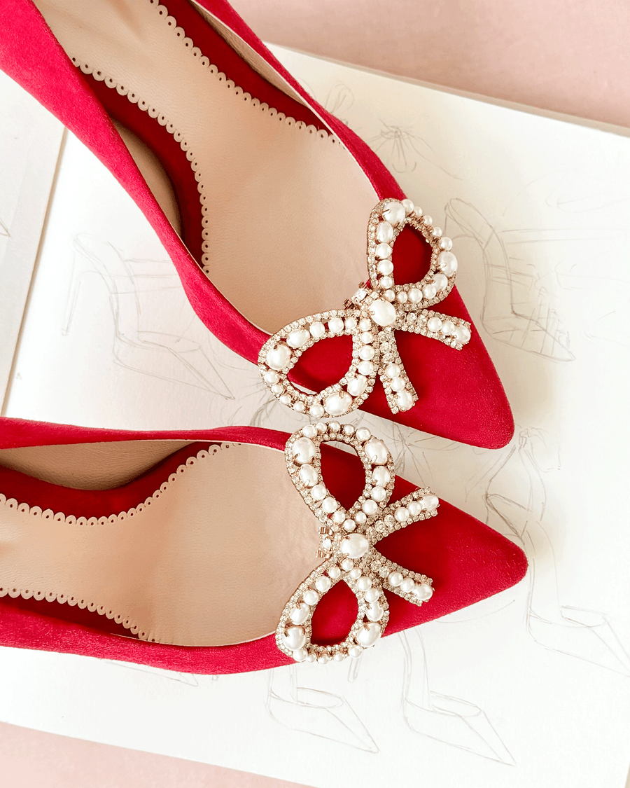 Red suede heels featuring large decorative bows adorned with pearls. The shoes are positioned on a light pink background with a sketch of a shoe design underneath, showcasing a top-down view. image