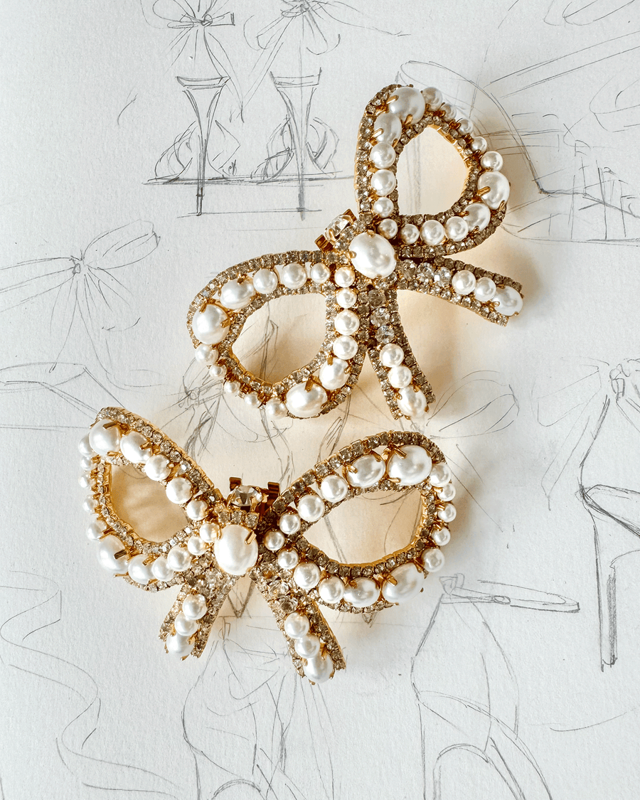 Two elegant hair clips designed in the shape of bows, adorned with pearls and sparkling rhinestones. The clips are set against a soft, textured background featuring fashion sketches, enhancing their luxurious appearance. The image is taken from a top-down angle, showcasing the intricate details of the clips. image