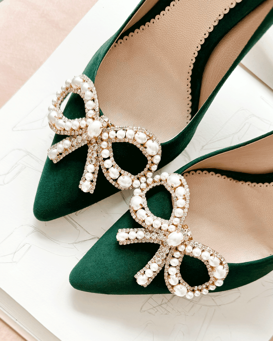 Green suede heels featuring a decorative bow adorned with pearls and rhinestones. The shoes are positioned on a light pink background, showcasing their pointed toe design. The image is taken from a top-down angle, highlighting the intricate details of the embellishments. image