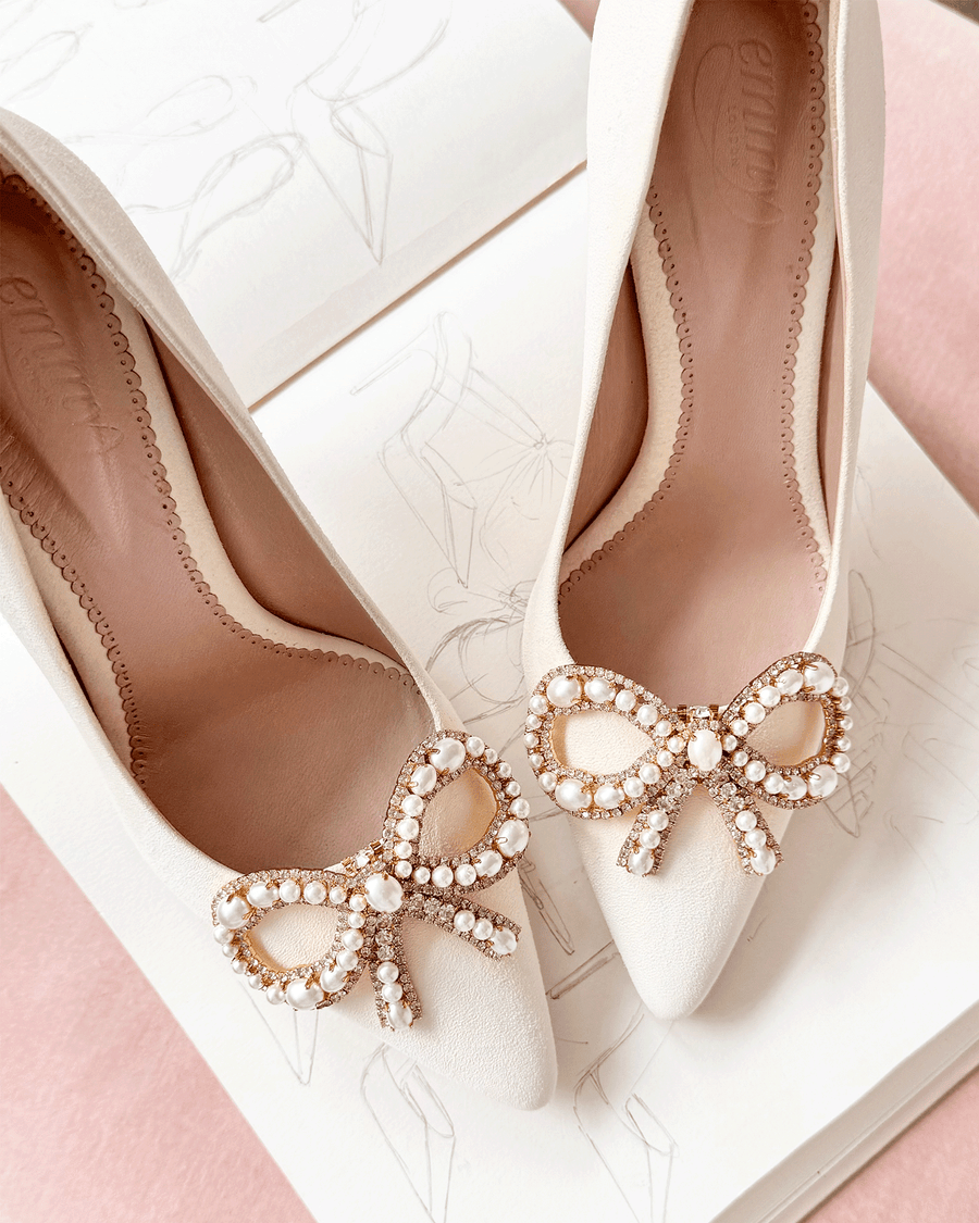 Elegant white heels featuring a decorative bow adorned with pearls and rhinestones at the toe. The shoes are positioned on a soft pink background, with a sketchbook displaying fashion illustrations in the background. The angle captures the shoes from above, highlighting their refined design and embellishments. image