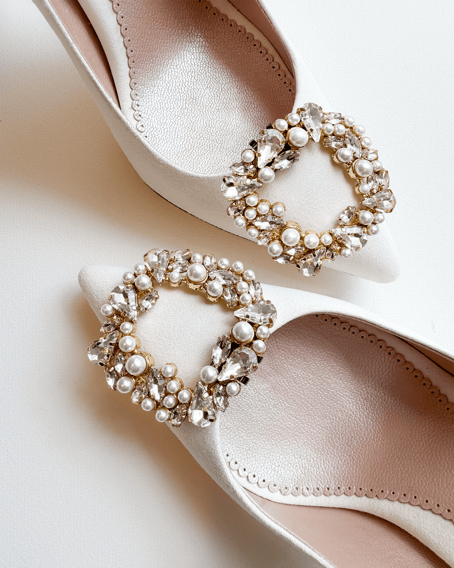 Elegant white heels featuring a decorative buckle adorned with pearls and crystals. The shoes are positioned on a neutral background, showcasing their pointed toe design and intricate embellishments. The angle highlights the details of the buckle and the smooth texture of the material. image
