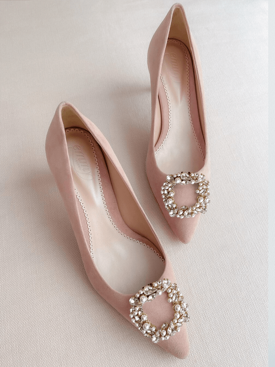 Elegant pink suede heels featuring a decorative crystal buckle on the toe. The shoes are displayed on a soft, neutral background, showcasing their pointed toe design. The image captures the shoes from a top-down angle, highlighting their sleek silhouette. image