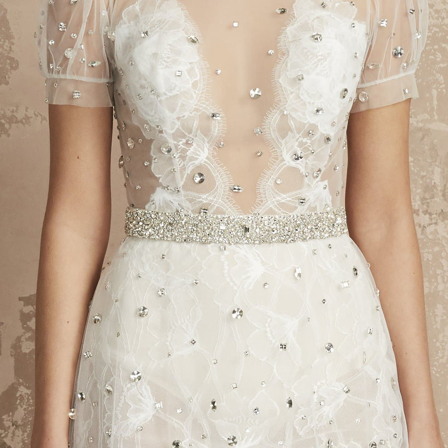 Elegant white dress featuring intricate floral lace and embellishments, with a sheer bodice and short puff sleeves. The dress is cinched at the waist with a sparkling crystal belt, set against a textured beige background. No shoes are visible in the image. image