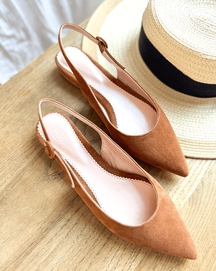Brown suede slingback heels are displayed on a wooden surface next to a straw hat. The shoes feature a pointed toe and a delicate ankle strap. The image is taken from a top-down angle, showcasing the smooth interior and the elegant design of the heels. image