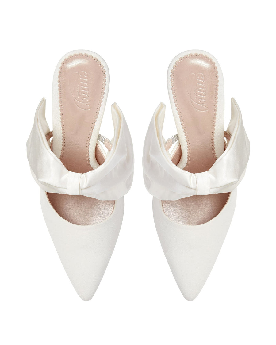 White pointed-toe mules featuring a satin bow detail at the back. The shoes have a sleek design with a smooth finish. The image is taken from a top-down angle against a plain white background, showcasing the elegant silhouette of the mules. image