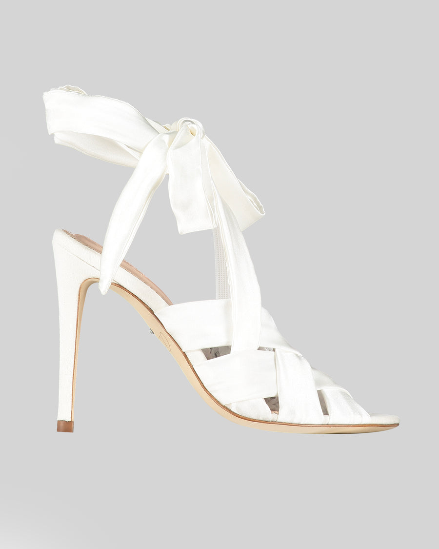 White high-heeled sandals featuring a strappy design with a ribbon tie at the ankle. The heel is approximately 4 inches tall, and the material is satin. The shoe is displayed against a light gray background, showcasing its elegant silhouette from a side angle. image