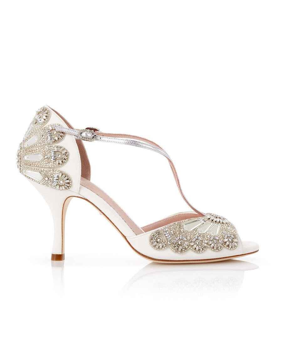 Elegant white heels featuring intricate beading and a T-strap design. The shoes have a slender heel, approximately 3 inches in height, and are made of leather. The image is taken from a side angle, showcasing the detailed embellishments and the delicate silhouette against a clean, neutral background. image