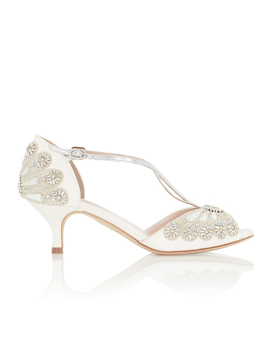 Elegant white heels featuring intricate beading and embellishments on the back. The shoes have a modest heel height, perfect for formal occasions. The image is taken from a side angle, showcasing the delicate straps and open toe design. The background is a soft, neutral tone, enhancing the shoe's details. image