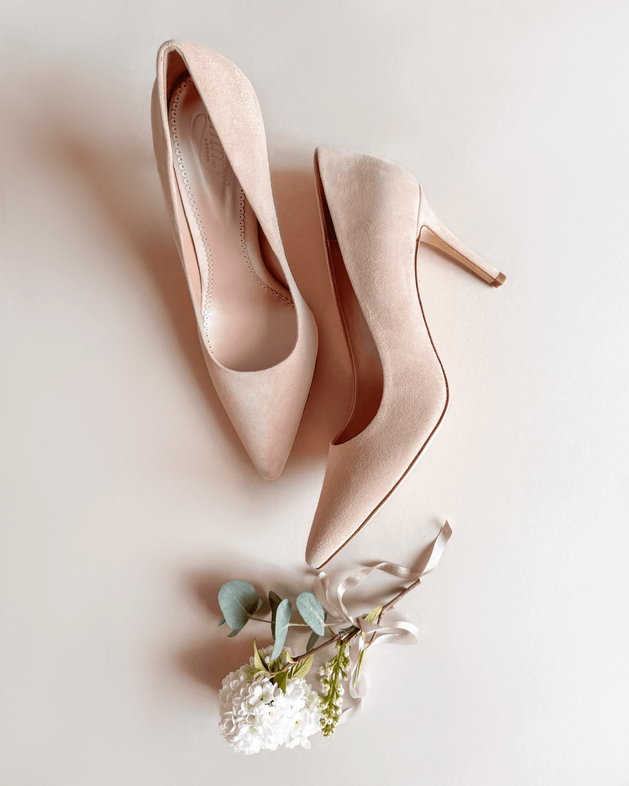 Nude suede heels with a pointed toe design, featuring a slim stiletto heel approximately 3 inches high. The shoes are positioned on a light beige background, accompanied by a delicate arrangement of greenery and a white flower, enhancing the elegant aesthetic. image