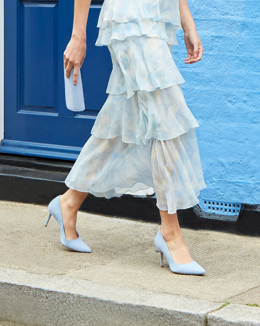 Light blue leather heels with a pointed toe, featuring a stiletto heel, are worn by a woman walking on a cobblestone street. She is dressed in a flowing, layered light blue dress, and is holding a matching clutch. The background showcases a vibrant blue wall, adding a pop of color to the scene. image