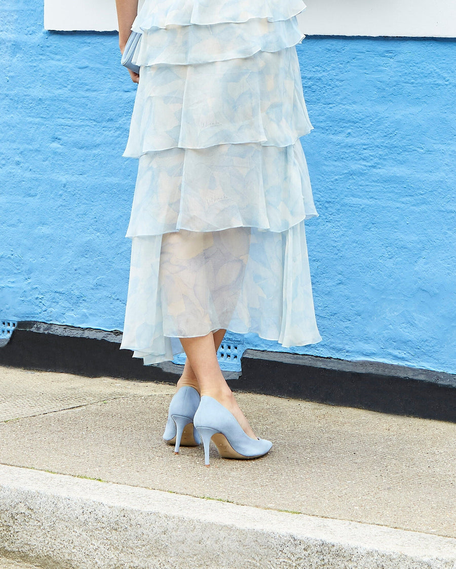 Light blue leather heels with a pointed toe, featuring a stiletto heel. The model is standing on a concrete surface, wearing a layered, semi-transparent dress that flows to mid-calf. The background is a vibrant blue wall, enhancing the overall aesthetic of the image. image