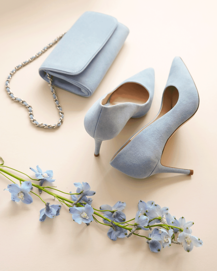 Light blue suede heels with a pointed toe and a stiletto heel are positioned next to a matching light blue clutch bag. The background is a soft, neutral color, and delicate blue flowers are arranged in front of the shoes, enhancing the overall aesthetic. The image is taken from a top-down angle, showcasing the elegant design of the heels. image