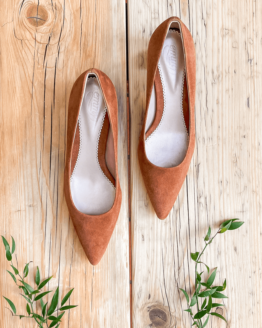 Brown suede pointed-toe heels are displayed on a rustic wooden surface, surrounded by small green leaves. The shoes are positioned from a top-down angle, showcasing their sleek design and soft texture. image