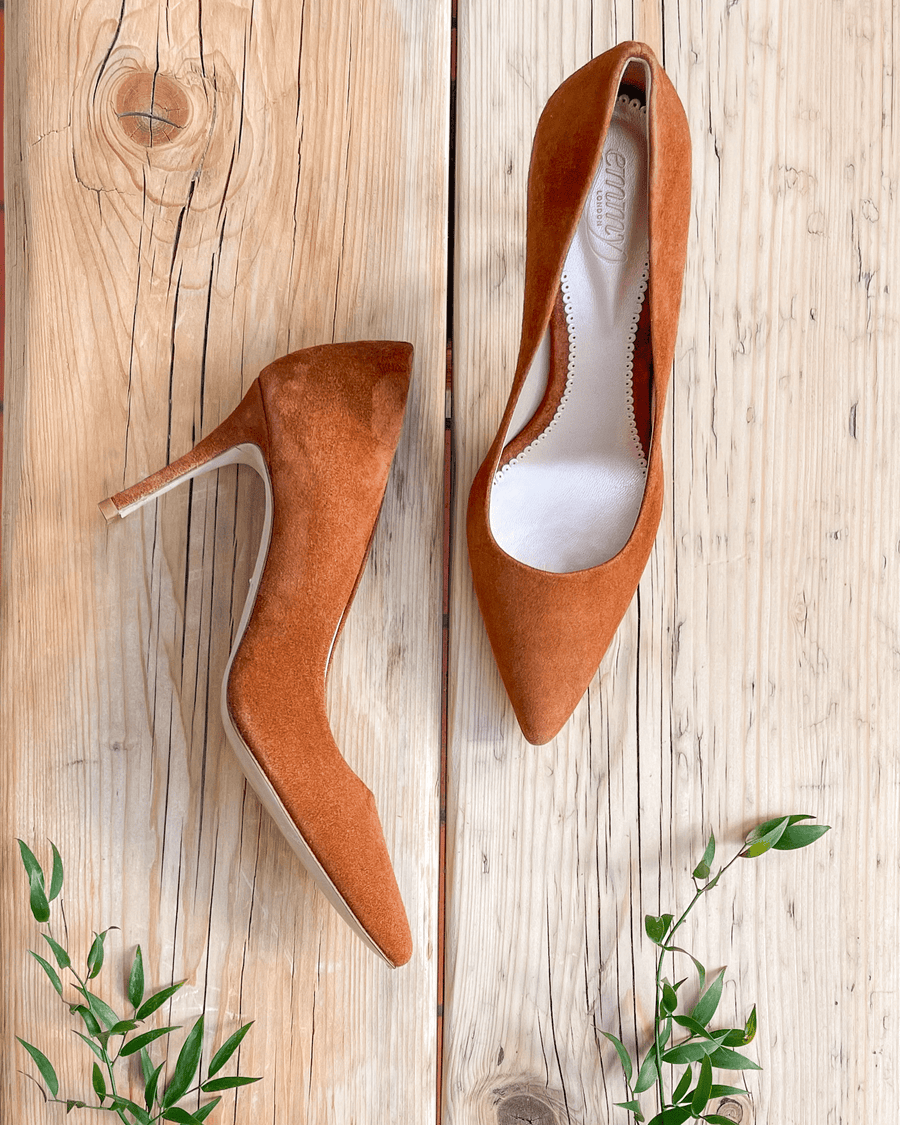Elegant orange suede heels are showcased on a rustic wooden surface, surrounded by delicate green foliage. The shoes feature a pointed toe and a stiletto heel, which measures approximately 4 inches. The image is taken from a top-down angle, highlighting the smooth texture of the suede and the clean lines of the design. image