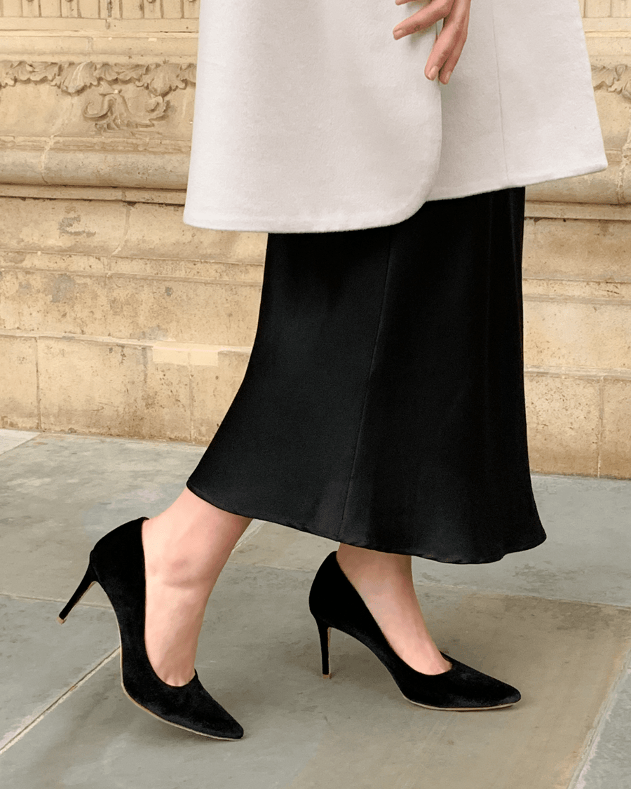Black suede heels with a pointed toe are worn by a person in a long black skirt, standing on a stone surface. The background features an ornate architectural detail, enhancing the elegant look of the outfit. The angle captures the shoes from the side, showcasing their height and style. image
