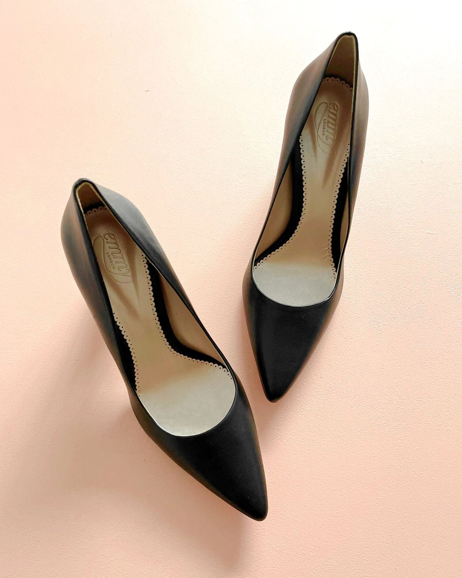 Black leather pointed-toe heels are displayed on a soft pink background. The shoes feature a sleek design with a smooth finish and a classic silhouette. The image captures the shoes from a top-down angle, showcasing their elegant shape and interior detailing. image