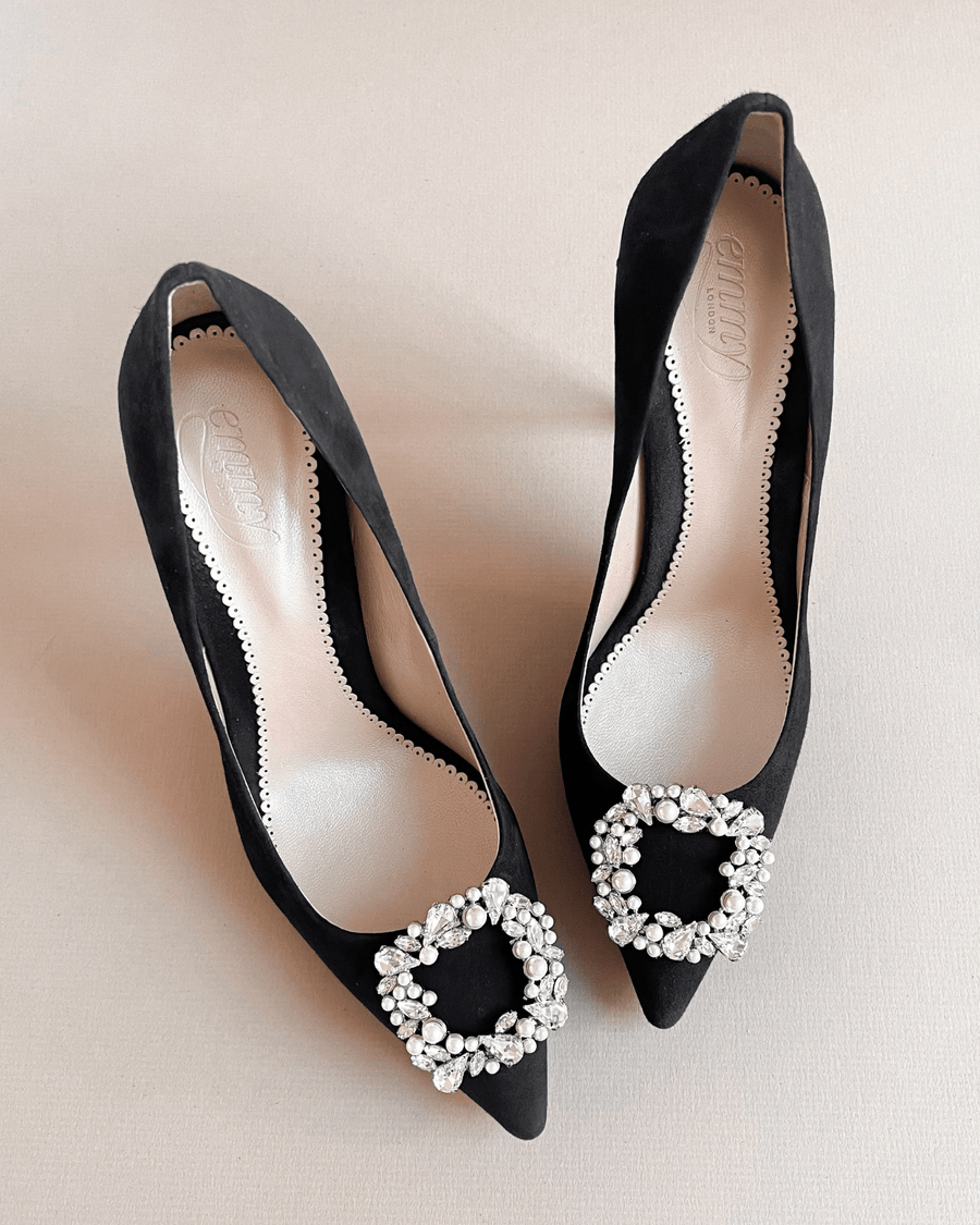 Black suede heels featuring a pointed toe design and embellished with a decorative crystal buckle on the front. The shoes are displayed on a light-colored background, showcasing their elegant silhouette. image