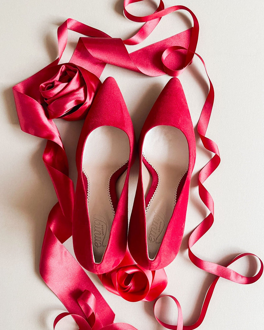Red suede heels are displayed on a light background, surrounded by pink satin ribbons and a rose. The shoes feature a pointed toe design and a sleek silhouette, emphasizing their elegant style. image