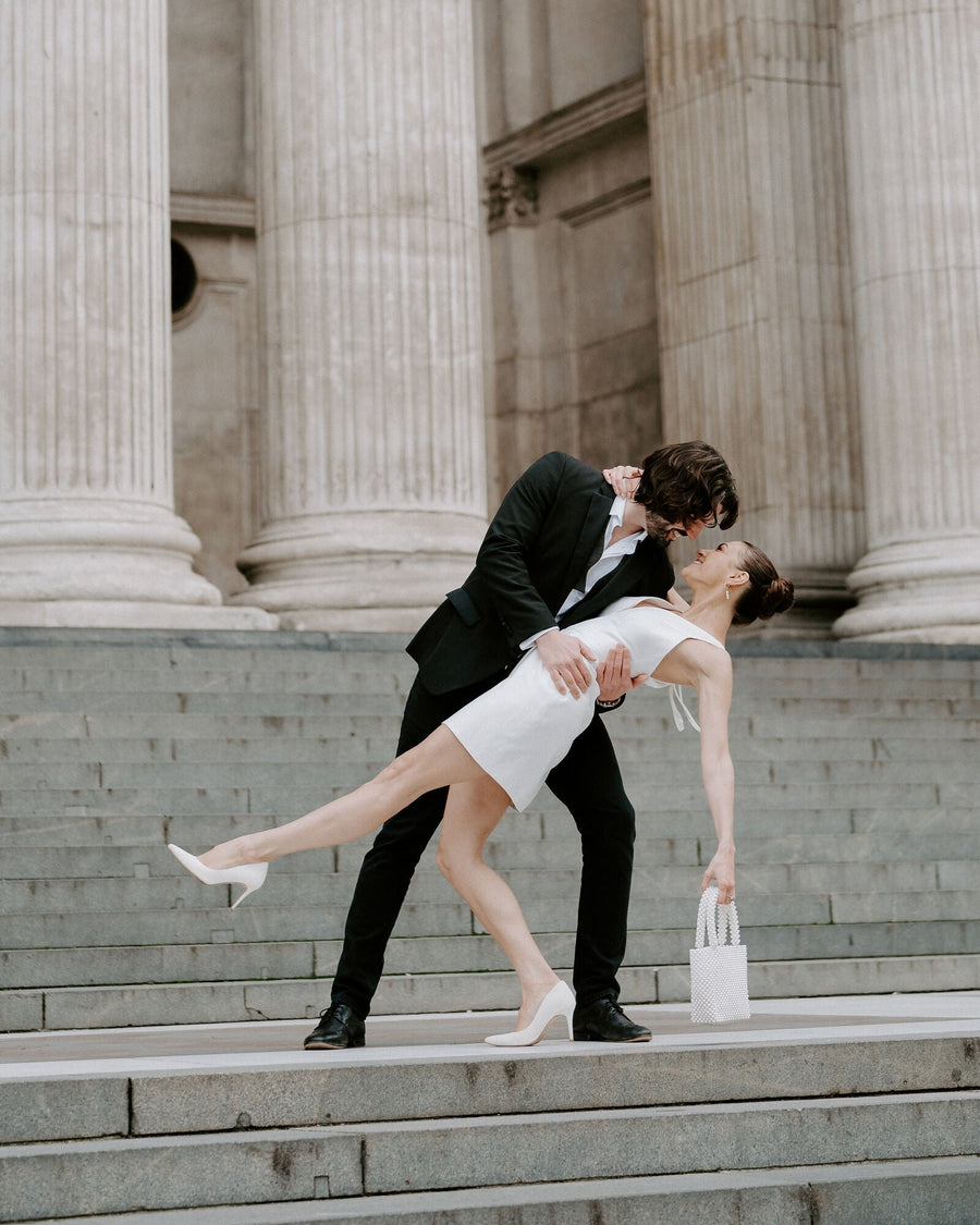 A couple is captured in a romantic pose on the steps of a grand building with tall columns. The woman is wearing a sleek white dress and white heels, while the man is dressed in a black suit. The image is taken from a low angle, emphasizing their elegant stance and the architectural details in the background. The woman is holding a small white handbag, and her heel height is approximately 4 inches. The heels are made of leather. image