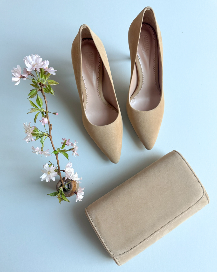 Nude suede high heels with a pointed toe design, positioned on a light blue background. A delicate branch with pink flowers is placed beside the shoes, and a matching beige clutch is positioned in front of them. The angle showcases the shoes from above, highlighting their sleek silhouette. image