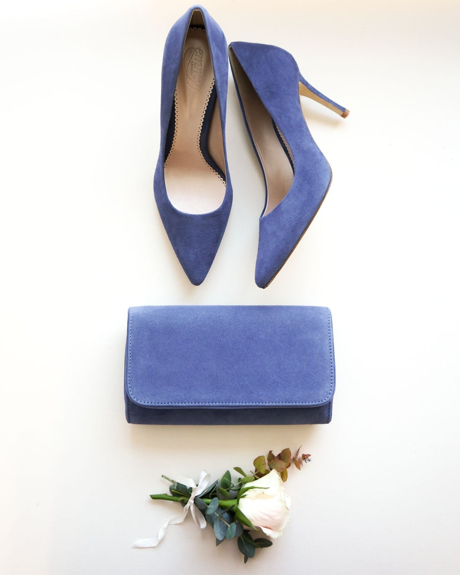 Blue suede heels with a pointed toe design are displayed alongside a matching blue clutch. The shoes feature a stiletto heel, positioned on a light background. A white rose and greenery are placed at the bottom of the image, adding a touch of elegance. image