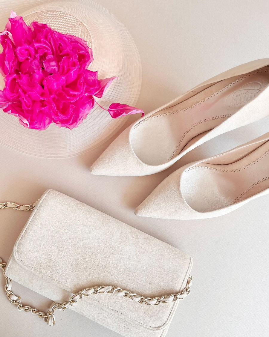 Elegant beige suede heels with a pointed toe design are positioned next to a chic clutch bag. The background features a soft, neutral surface, and a vibrant pink flower adds a pop of color to the scene. The shoes are displayed from a top-down angle, showcasing their sleek silhouette. image