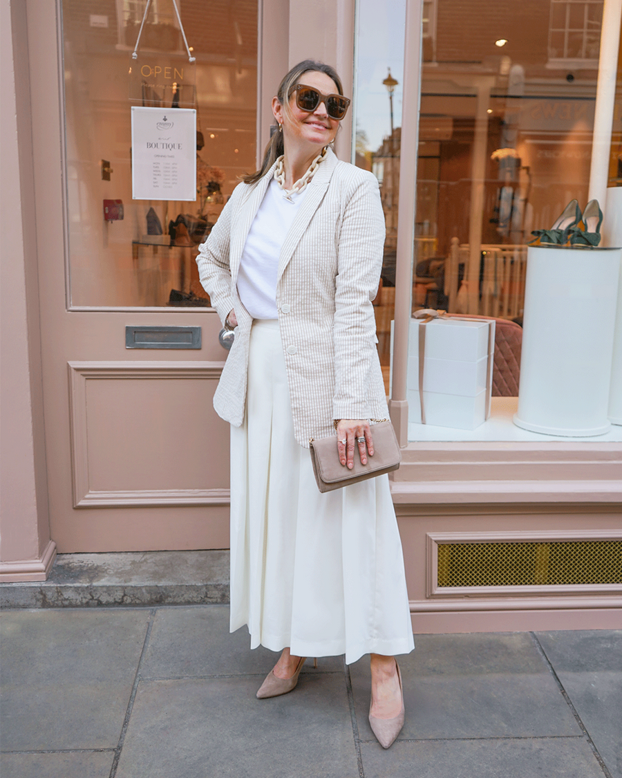 A woman stands outside a boutique, wearing a stylish cream blazer over a white top and a flowing white skirt. She accessorizes with sunglasses and holds a beige handbag. The heels are a light taupe color, complementing her outfit. The image is taken from a side angle, showcasing the boutique's window display in the background. image