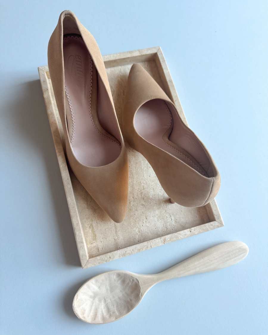 Beige suede heels with a pointed toe design are displayed on a light-colored tray. The shoes are positioned at an angle, showcasing their sleek silhouette. A wooden spoon is placed beside the tray, adding a natural element to the composition. The background is minimalistic, emphasizing the shoes. image
