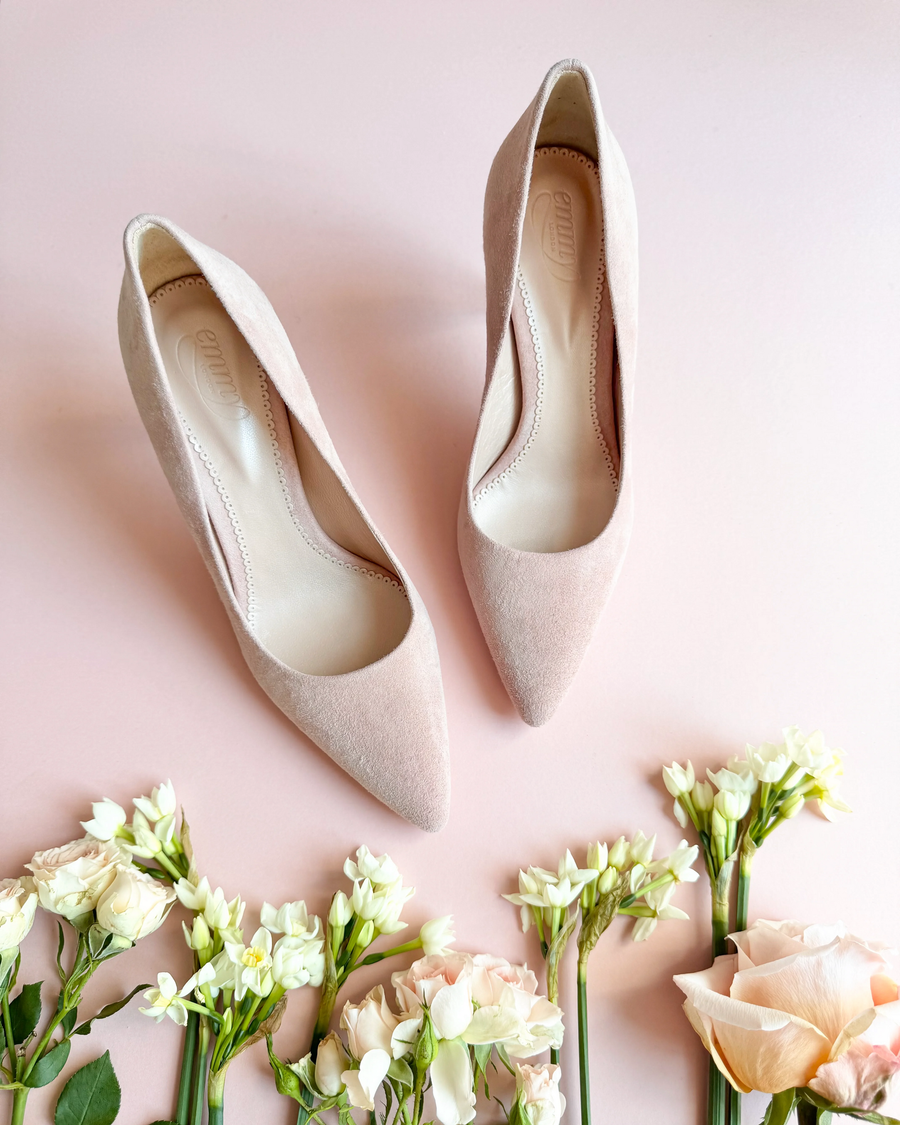 Elegant blush suede heels are showcased against a soft pink background, surrounded by delicate white and pastel flowers. The shoes feature a pointed toe and a sleek silhouette, emphasizing their sophisticated design. image
