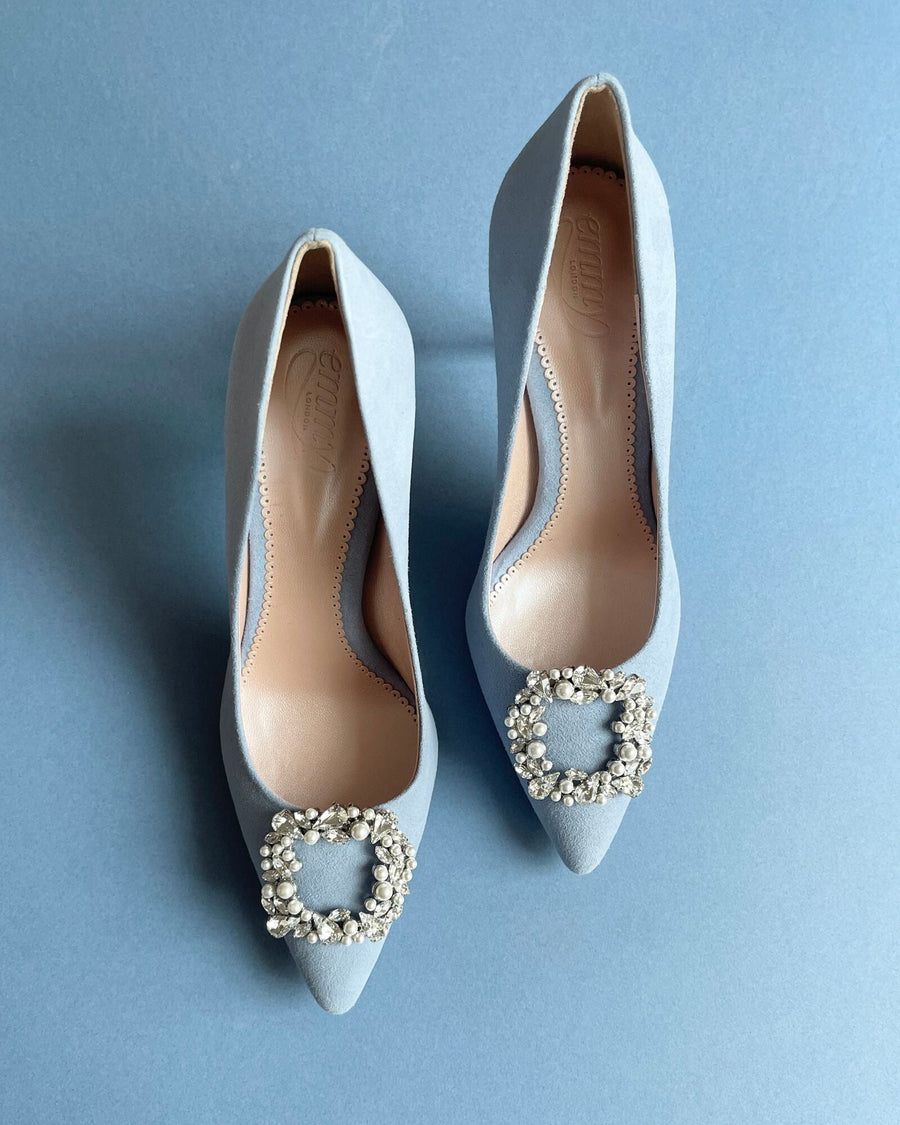 Light blue suede heels with a pointed toe and a decorative crystal buckle on the vamp. The shoes are displayed on a solid light blue background, showcasing their elegant design from a top-down angle. image