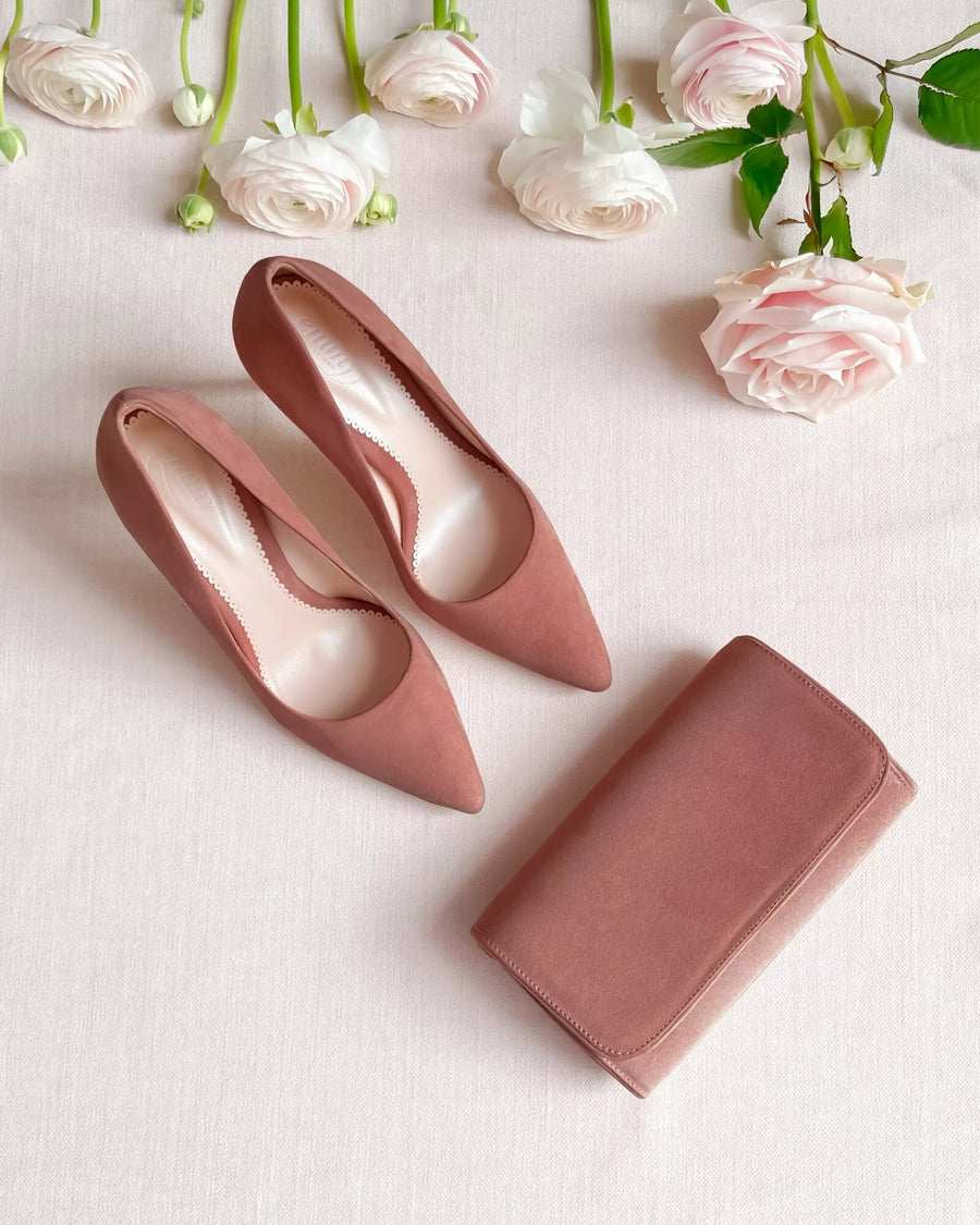 Elegant blush suede heels with a pointed toe design are displayed on a soft, neutral fabric background, accompanied by a matching clutch. The shoes are positioned side by side, showcasing their sleek silhouette, while delicate pink roses and greenery are arranged around them, adding a touch of romance to the scene. image