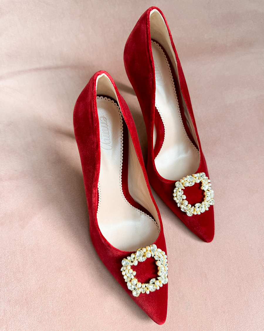 Red suede heels with a pointed toe and a decorative crystal buckle on the front. The shoes are displayed on a soft, blush-colored background, showcasing their elegant design. The angle captures both shoes from above, emphasizing their sleek silhouette. image