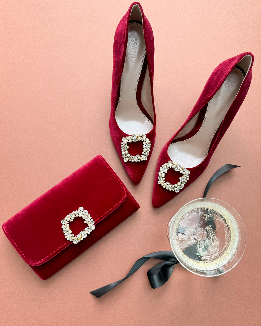 Red suede high heels with a pointed toe and embellished buckle detail, positioned alongside a matching red clutch and a glass of champagne on a soft pink background. The shoes are displayed from a top-down angle, showcasing their elegant design. image