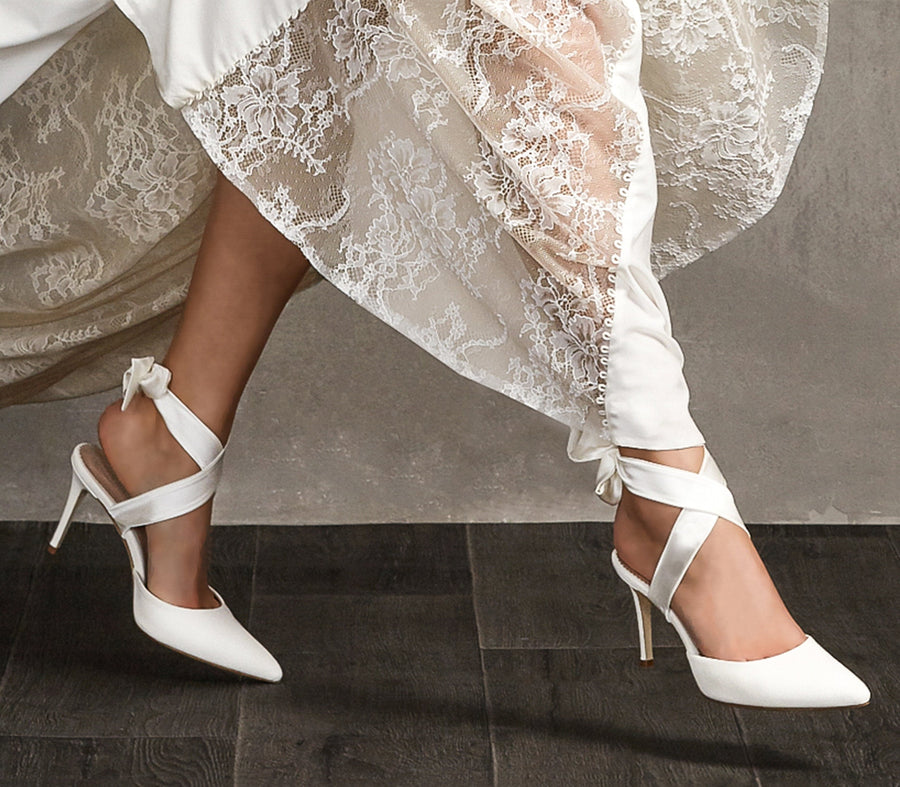 White stiletto heels with a pointed toe and ankle straps, featuring a sleek satin finish. The model is wearing them with a flowing, lace-trimmed dress, captured from a side angle on a dark wooden floor. The heel height is approximately 4 inches. image