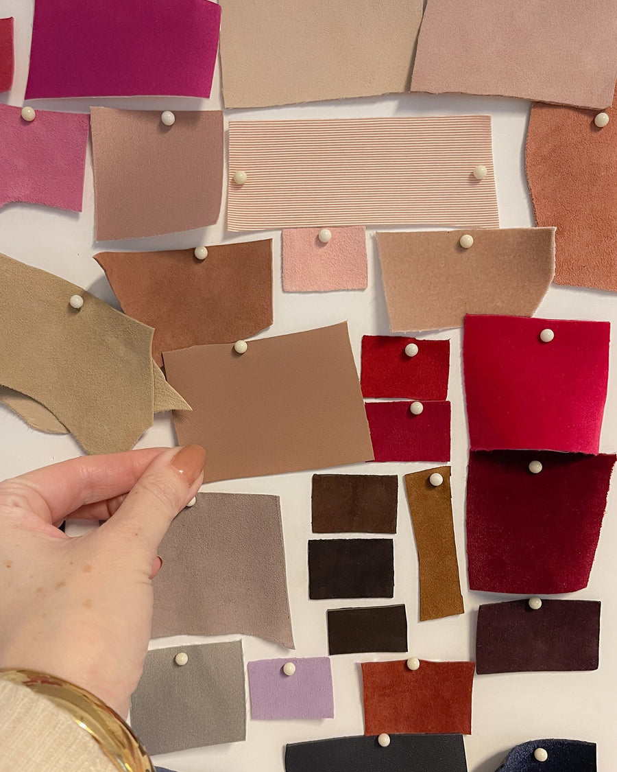 The image shows a hand holding a brown swatch of material against a backdrop of various fabric samples pinned to a wall. The samples include a range of colors and textures, predominantly in shades of pink, beige, and burgundy. The angle captures the hand in the foreground, emphasizing the swatch while the colorful fabric pieces create a visually appealing collage in the background. image