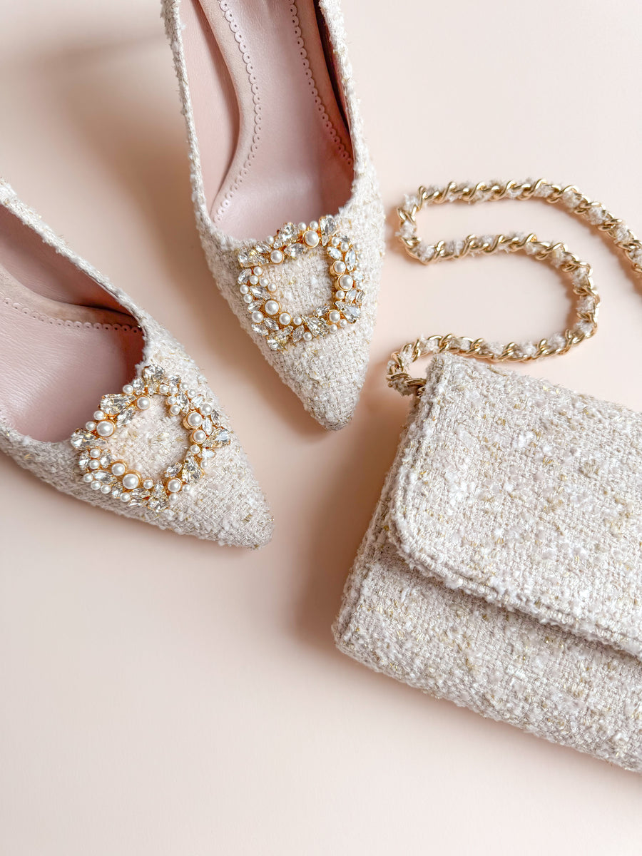 Elegant tweed heels with a pointed toe design, featuring a decorative pearl buckle on the front. The shoes are positioned on a soft, neutral background alongside a matching tweed handbag with a gold chain strap. The image captures the shoes from a top-down angle, showcasing their intricate texture and detailing. image
