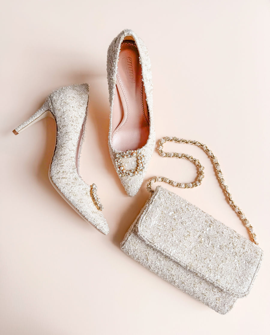 Elegant tweed heels with a pointed toe and a decorative embellishment on the front, accompanied by a matching clutch bag. The shoes are positioned on a soft, neutral background, showcasing their intricate texture and design. The image captures the heels from a side angle, highlighting their height and stylish silhouette. image