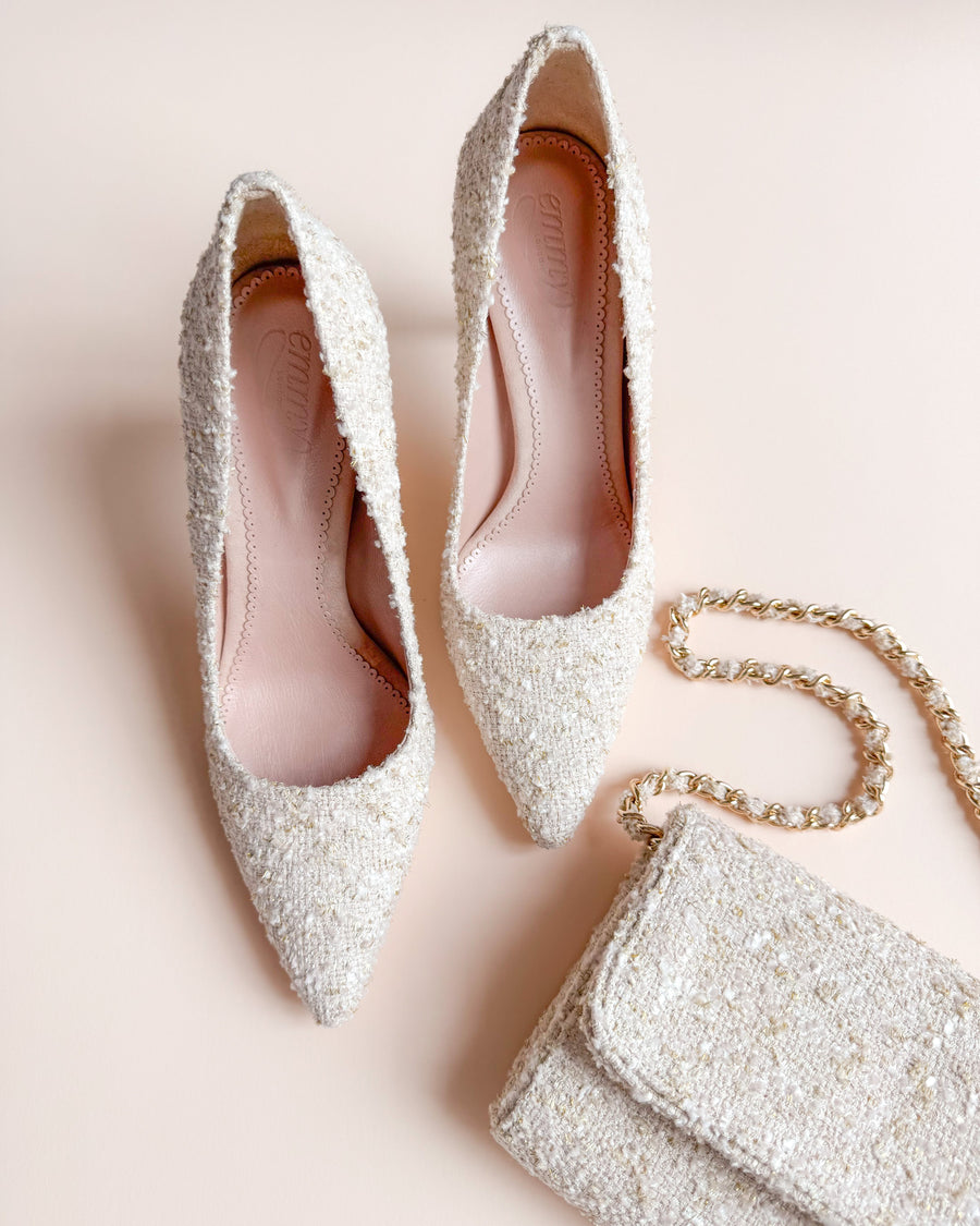 Elegant pointed-toe heels in a textured cream tweed material, featuring a slim stiletto heel. The shoes are positioned on a soft, neutral background, accompanied by a matching small handbag with a chain strap, creating a chic ensemble. image