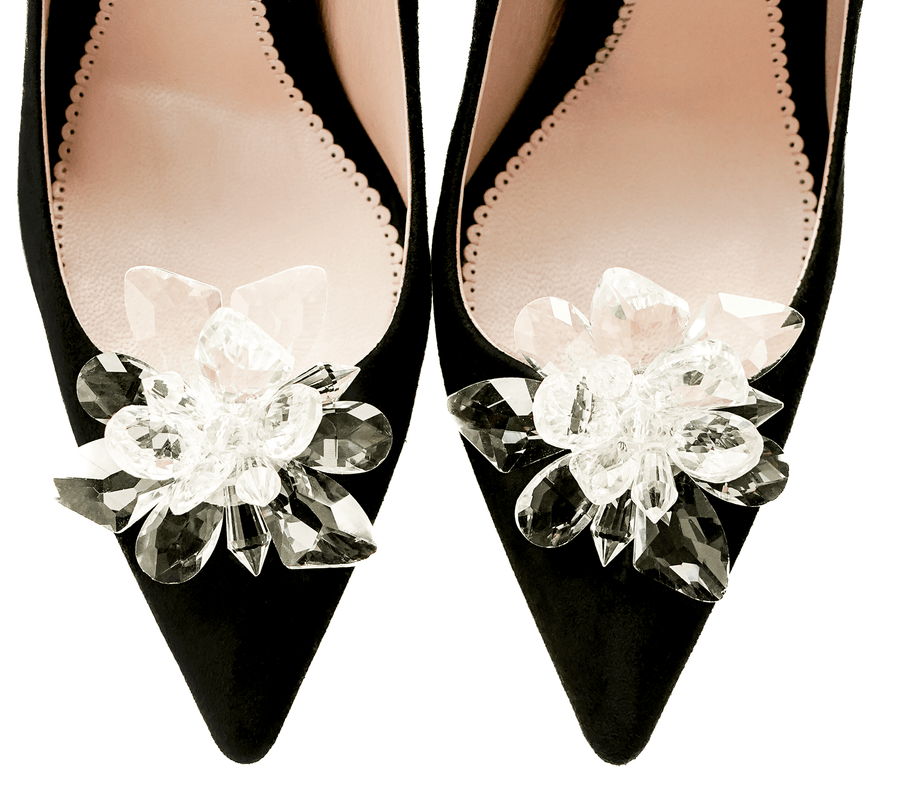 Black suede heels featuring a decorative crystal flower embellishment on the toe. The image showcases a top-down view of the shoes, highlighting the intricate details of the embellishment against a soft pink insole. The background is neutral, allowing the shoes to stand out prominently. image