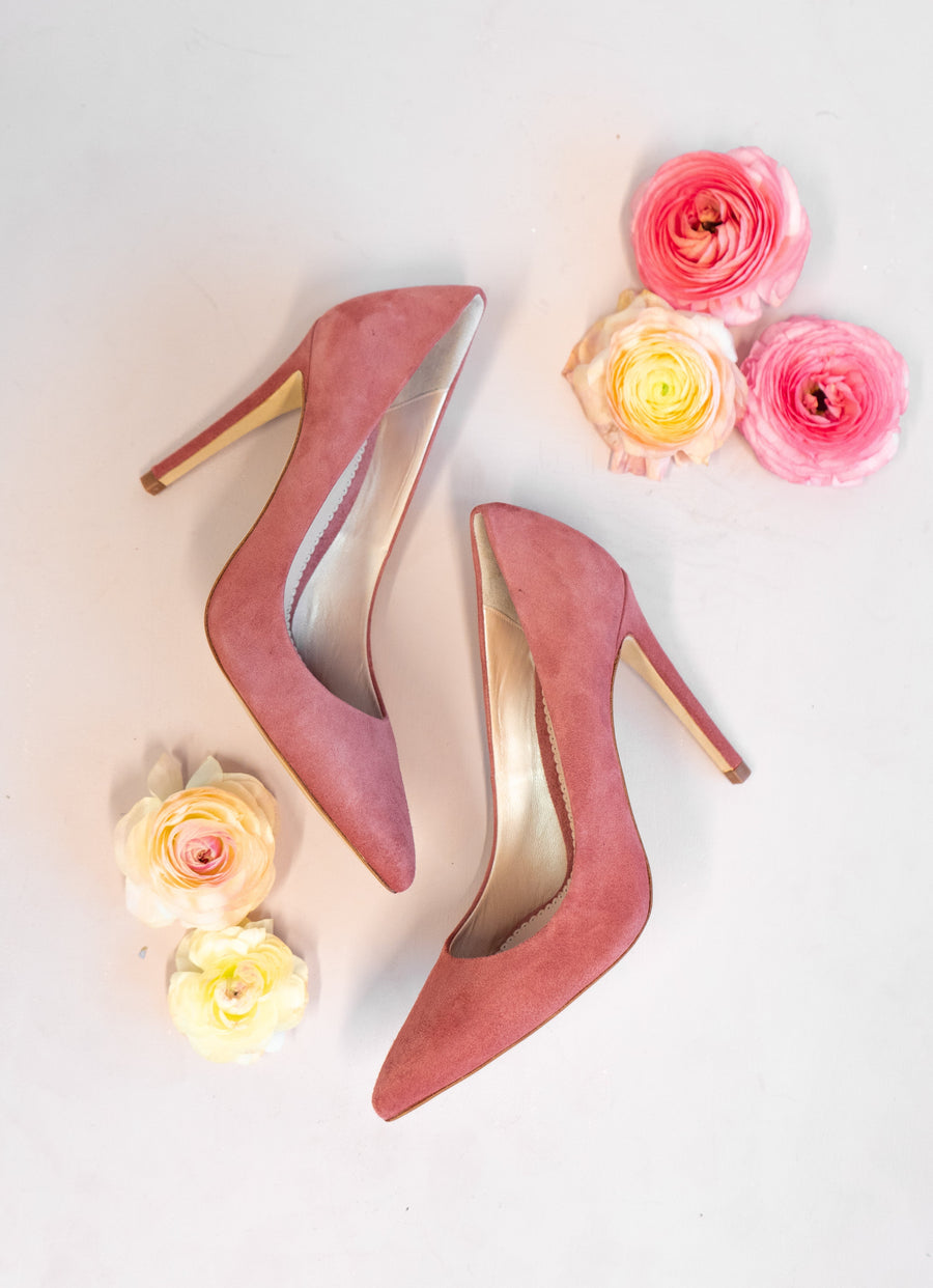Pink suede high heels are displayed on a light background, surrounded by soft pastel flowers. The shoes feature a pointed toe and a sleek silhouette, showcasing their elegant design. image