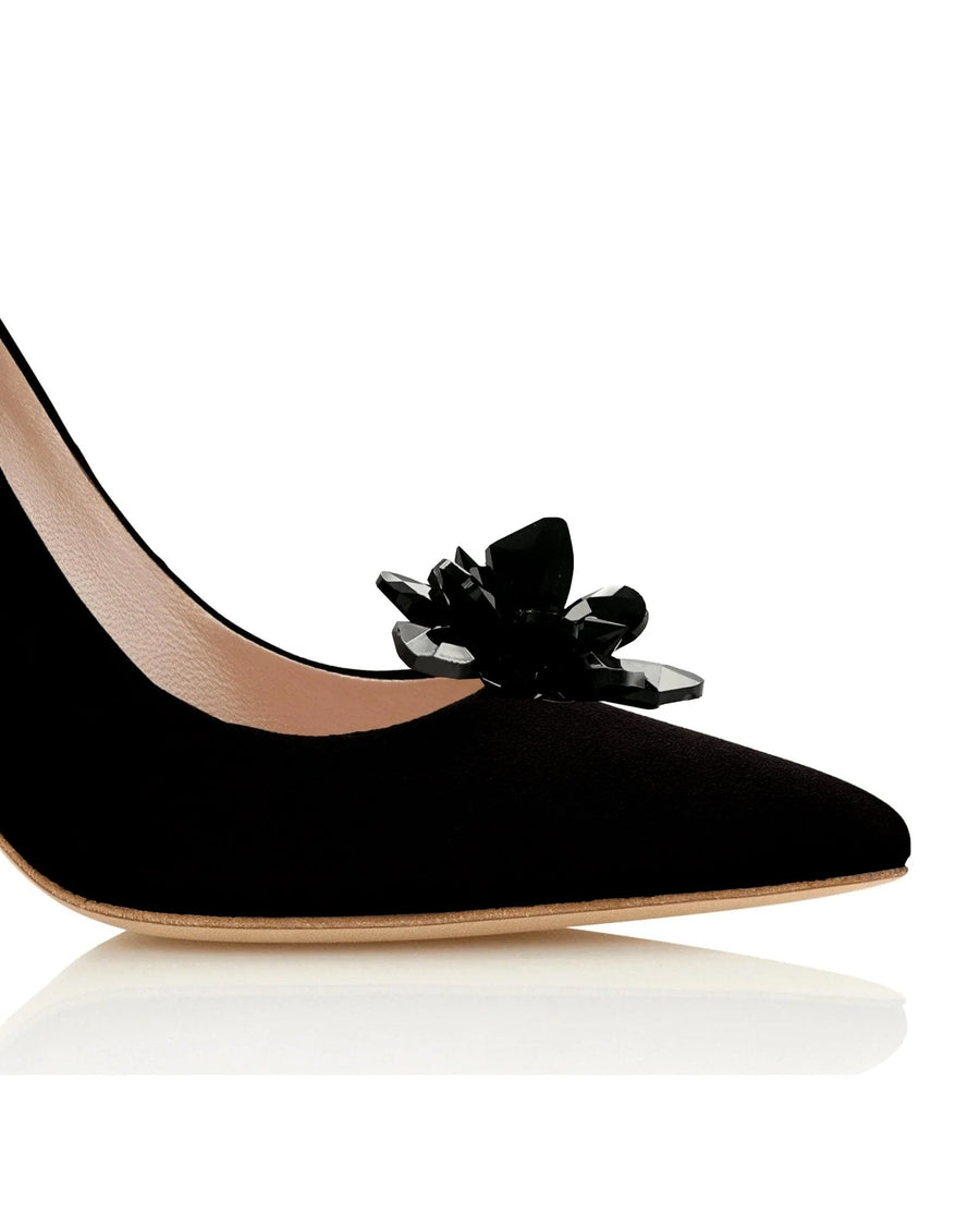 Black suede heels featuring a pointed toe and an elegant black floral embellishment on the vamp. The image is taken from a close-up angle, highlighting the shoe's sleek design and texture against a clean, white background. image