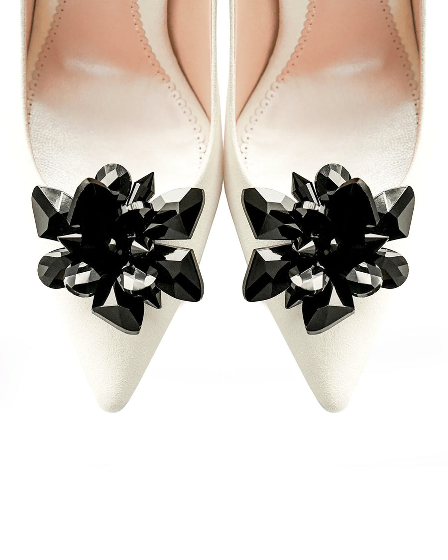 Elegant white pointed-toe heels adorned with a striking black crystal floral embellishment at the front. The shoes are displayed against a soft, neutral background, highlighting their sophisticated design. The angle captures the shoes from above, showcasing the intricate detailing of the embellishment. image