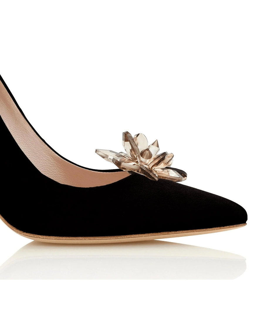 Black suede heels featuring a decorative crystal embellishment at the toe. The image is taken from a low angle, highlighting the pointed toe and sleek silhouette. The background is a clean, white surface, emphasizing the shoe's elegant design. image
