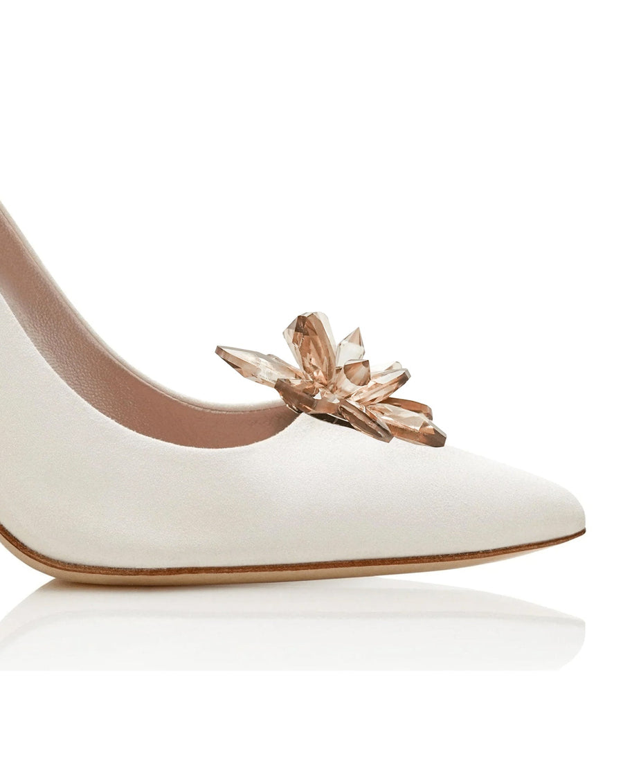 White suede heels featuring a prominent crystal embellishment on the toe. The image is taken from a side angle, showcasing the sleek silhouette and pointed toe design. The background is a clean, reflective surface that highlights the shoe's details. image